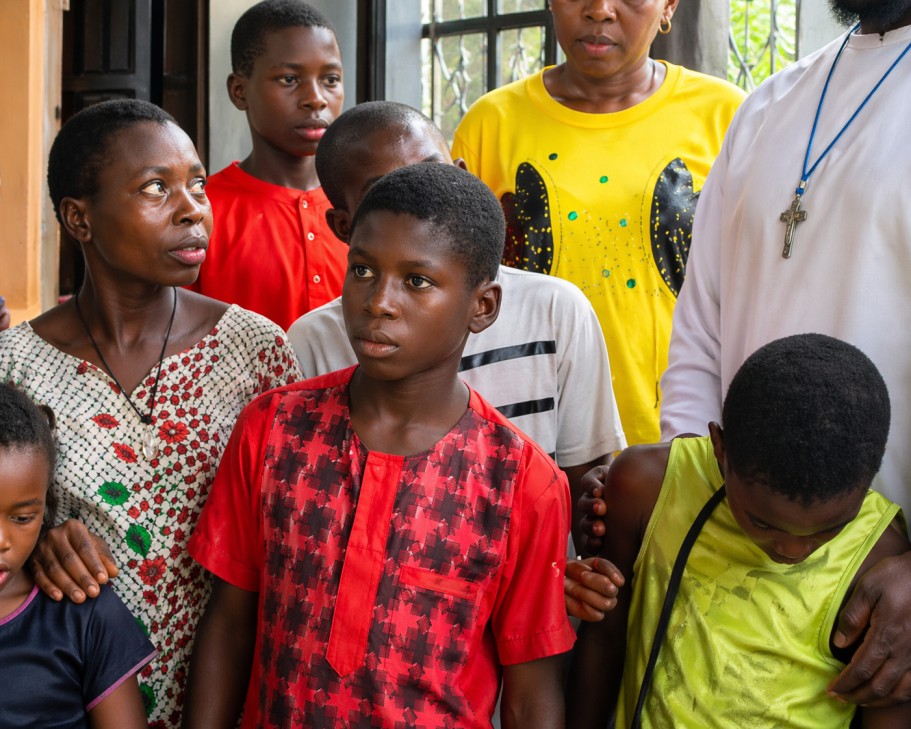 A community leader in white robes standing with a group of attentive youth and women during an outreach program.