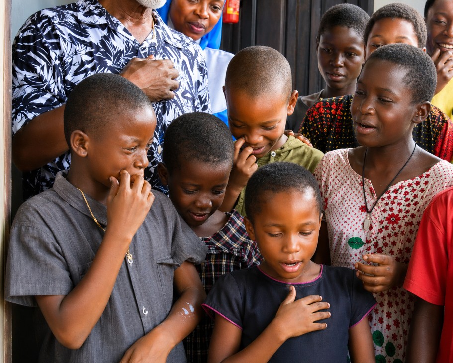 Diverse group of happy school-aged children in colorful clothing gathered around a community leader in a bright outdoor setting.